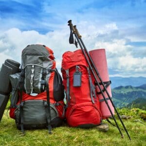 Two red backpacks with hiking poles and sleeping mats on a grassy hilltop, overlooking a mountainous landscape under a partly cloudy sky.