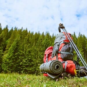 A loaded backpack, trekking poles, and a rolled-up mat are on grass with a forest backdrop under a partly cloudy sky.