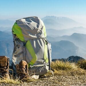 A backpack and hiking boots on a grassy hilltop with mountains and a hazy sky in the background.